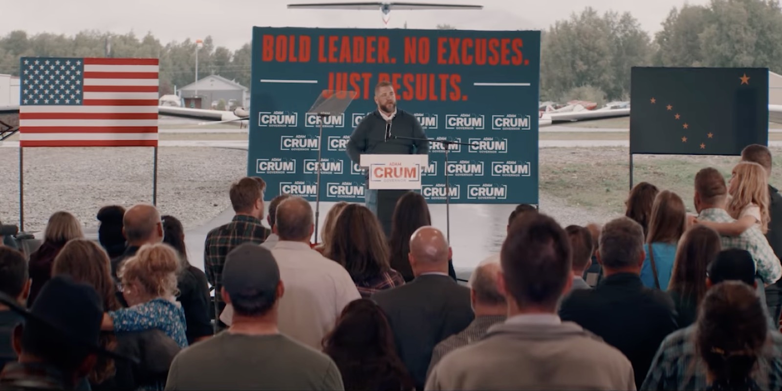 Adam Crum speaking from a podium to a seated crowd at an Alaska campaign rally, under a banner reading Bold Leader. No Excuses. Just Results.